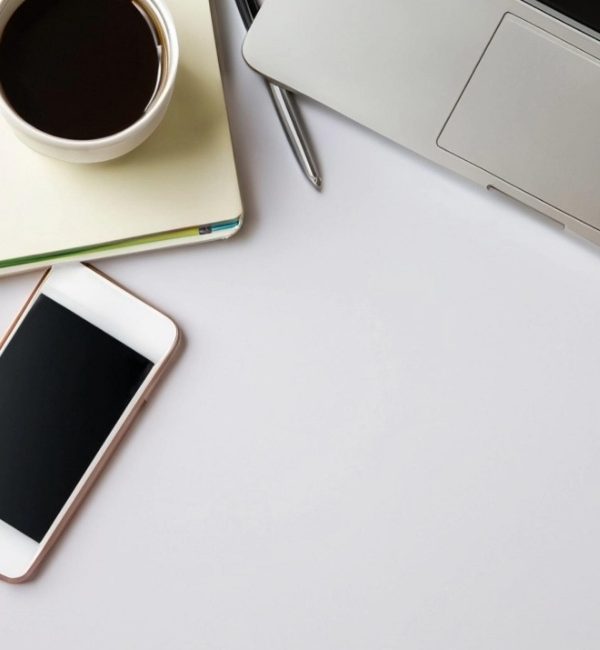 Top view of a laptop, mobile phone, coffee cup and notebook on a white desk